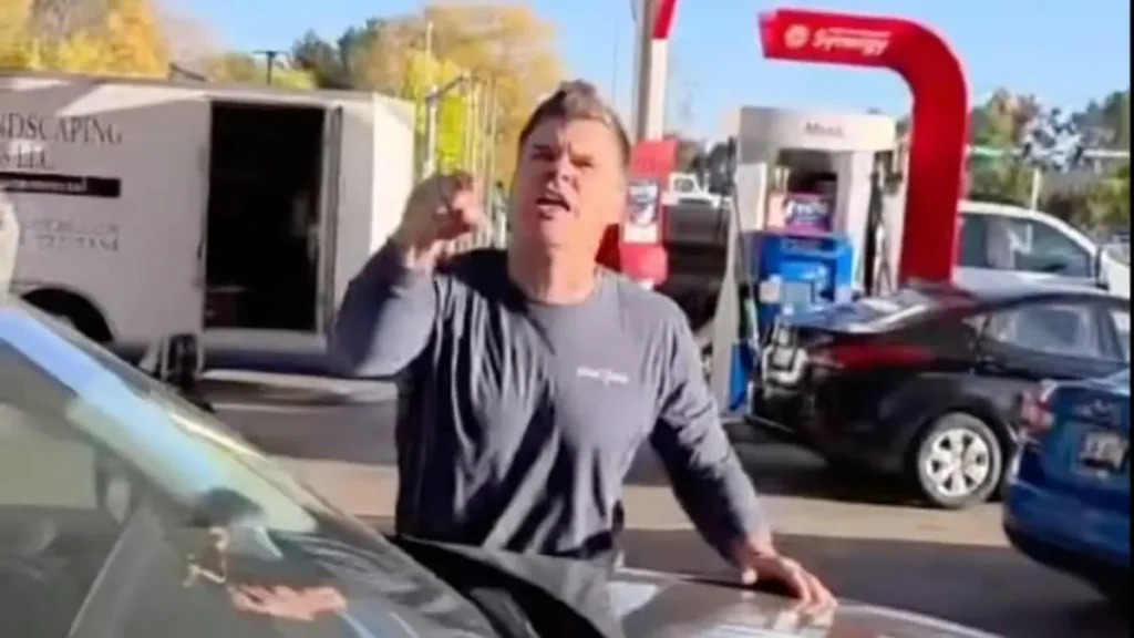 A man gestures emphatically while standing next to a car in a gas station parking lot.
