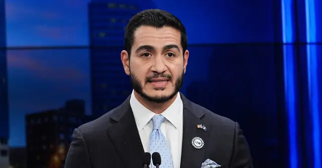 The image shows a man in a suit and tie, standing at a podium with microphones, in what appears to be a news studio setting.