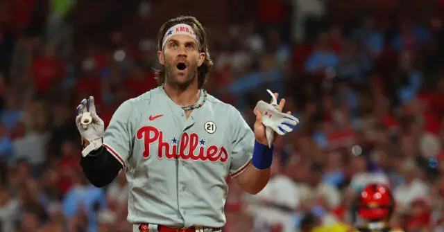 A baseball player in a Philadelphia Phillies uniform celebrates with a surprised expression and raised hands.