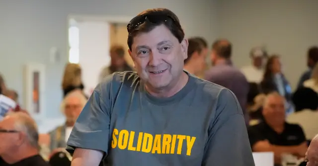 The photo shows a man wearing a shirt that says "SOLIDARITY" in a room with other people in the background.