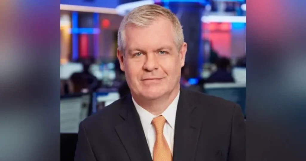 The image shows a news anchor in a professional setting, wearing a dark suit and an orange tie.