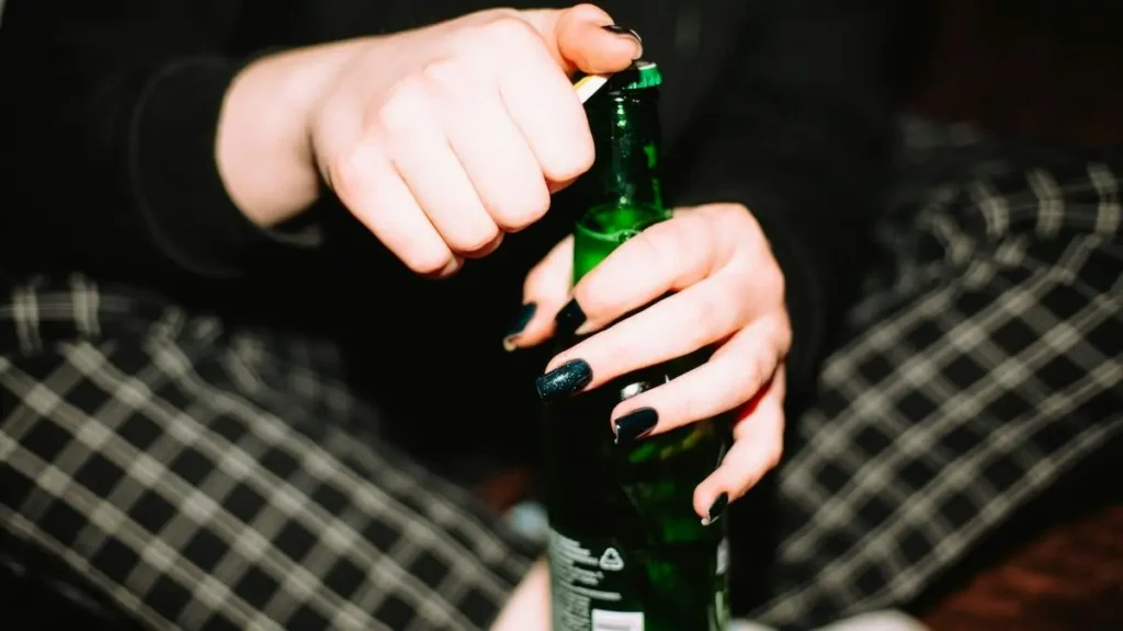A person with dark nail polish is opening a green bottle, possibly a beer, while sitting on a checkered surface.
