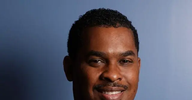 The image shows a close-up of a smiling individual with short, curly hair against a plain blue background.