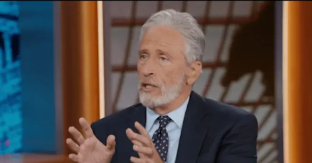 The image shows a man in a suit and tie, gesturing with his hands while speaking, likely during a news segment or interview.