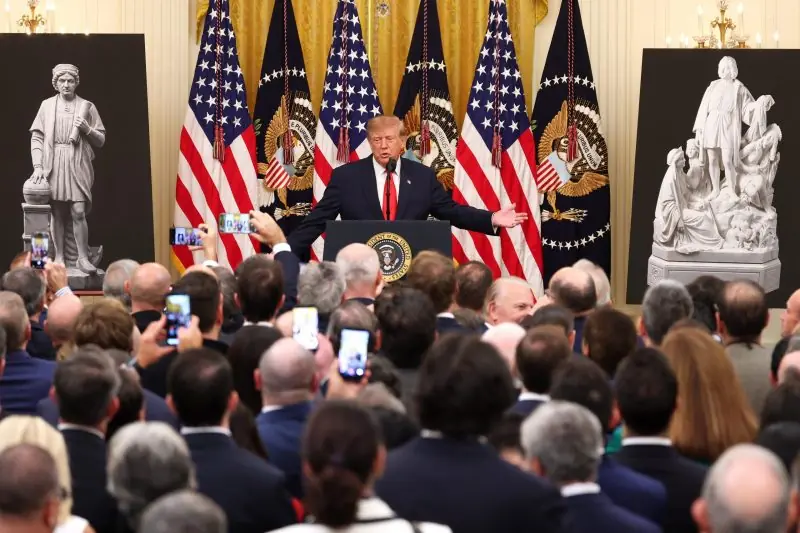 The image shows a man speaking at a podium in front of a large audience, with American flags and statues in the background.