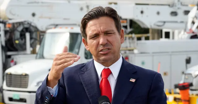 A man in a suit and red tie is speaking into a microphone in front of a large truck.