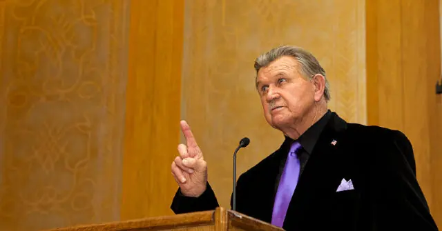 The image shows a man in a suit and tie giving a speech at a podium, gesturing with his right hand.