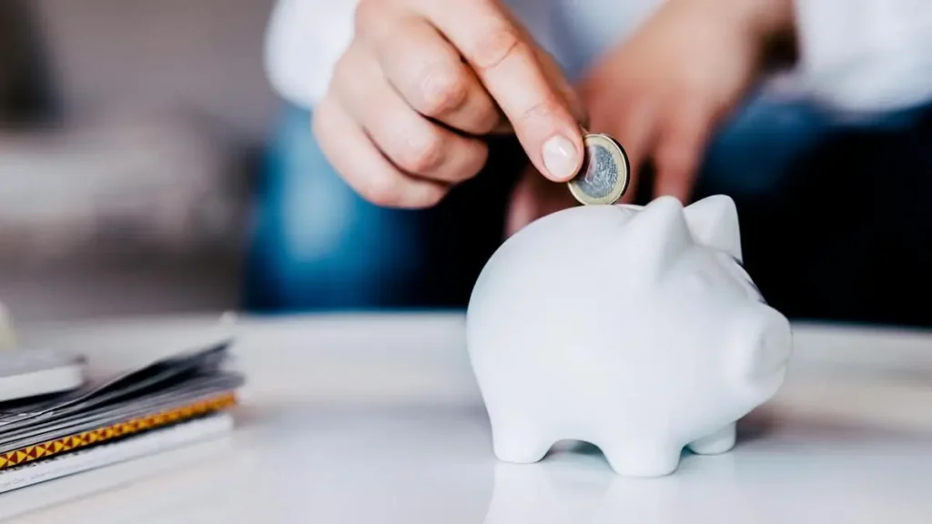 A person is placing a coin into a white piggy bank, symbolizing saving money.