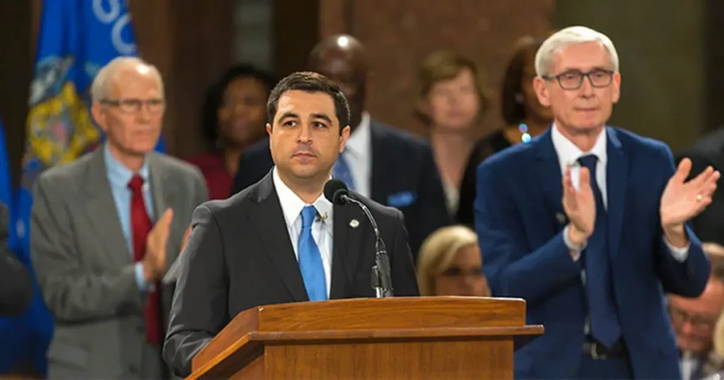 A man in a suit stands at a podium, delivering a speech, while others in the background applaud.