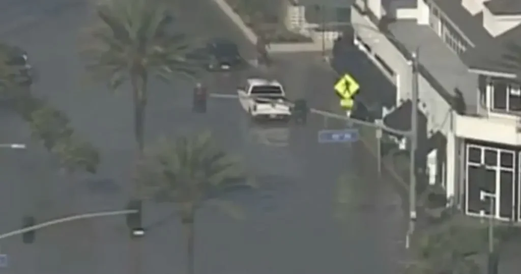 A vehicle drives through a flooded street with palm trees and buildings visible in the background.