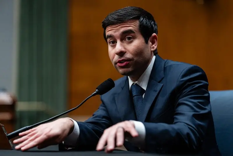 The image shows a man in a suit and tie speaking at a formal event, with a microphone in front of him.