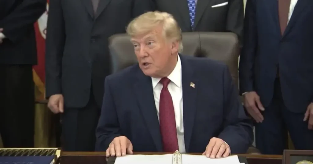 The image shows a formal setting where a person in a suit is seated at a desk, with others in suits standing behind, suggesting a significant official or ceremonial event.