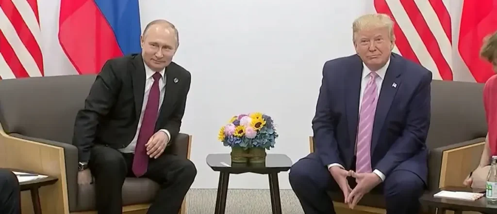 The image shows two individuals in formal attire seated in front of flags, with a small table in between them holding a bouquet of flowers.