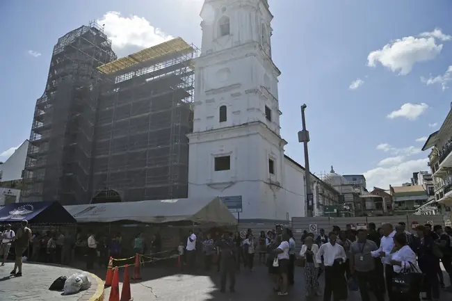 The photo shows a bustling public square with a historic white tower under construction, surrounded by scaffolding, and a crowd of people gathered nearby.