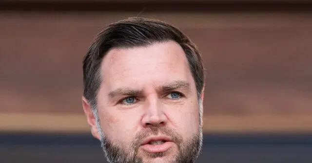 The image shows a close-up of a man with short dark hair and a beard, wearing a suit, against a blurred background.