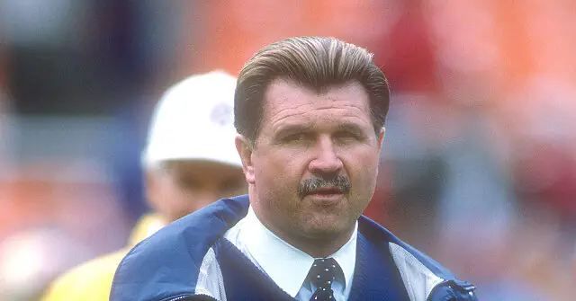 The image shows a man in a sports setting, wearing a jacket and tie, with a focused expression.