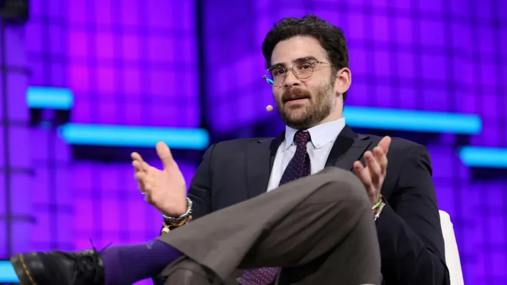 A man in a suit and tie is gesturing with his hands while speaking on stage in front of a purple-lit backdrop.