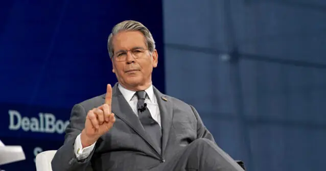 The image shows a man in a suit gesturing with his right hand while speaking at a formal event.
