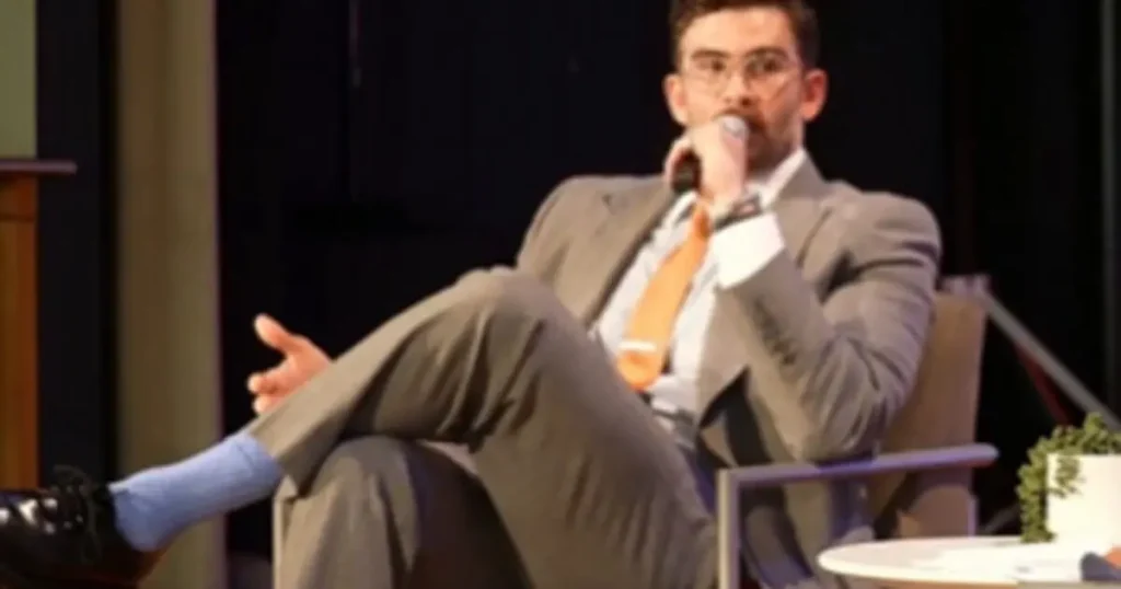 A man in a suit and tie is sitting back in a chair with his feet up, speaking into a microphone.