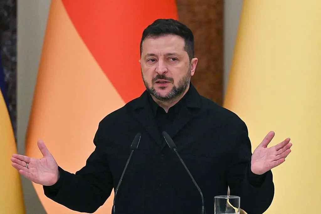 The image shows a person speaking at a podium with a microphone, with flags in the background, suggesting a formal or official setting.