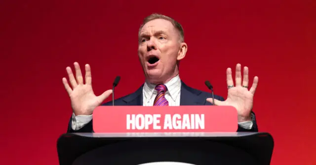 A man stands at a podium with a "HOPE AGAIN" sign, gesturing passionately with his hands raised.