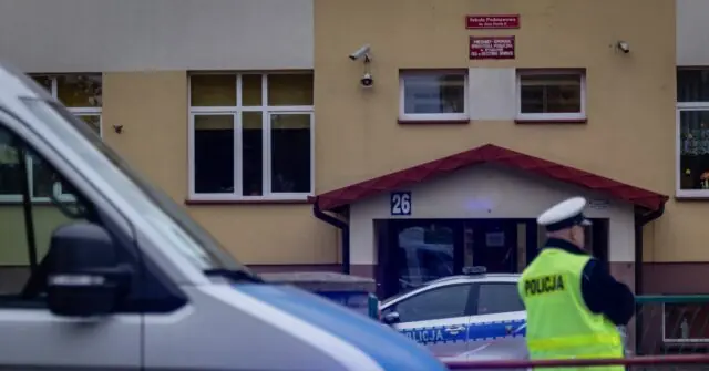 A police officer stands near a building with a "Policja" vest, indicating a law enforcement presence, possibly at a crime scene or security checkpoint.