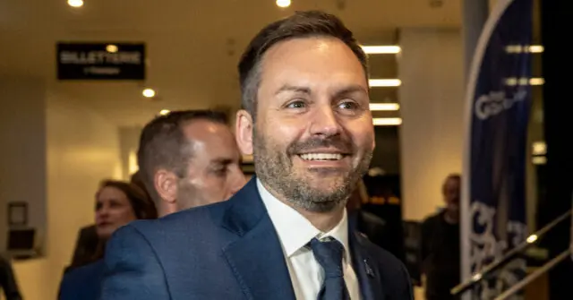 The image shows a man in a suit smiling in what appears to be a professional or formal setting, possibly at an event or conference.